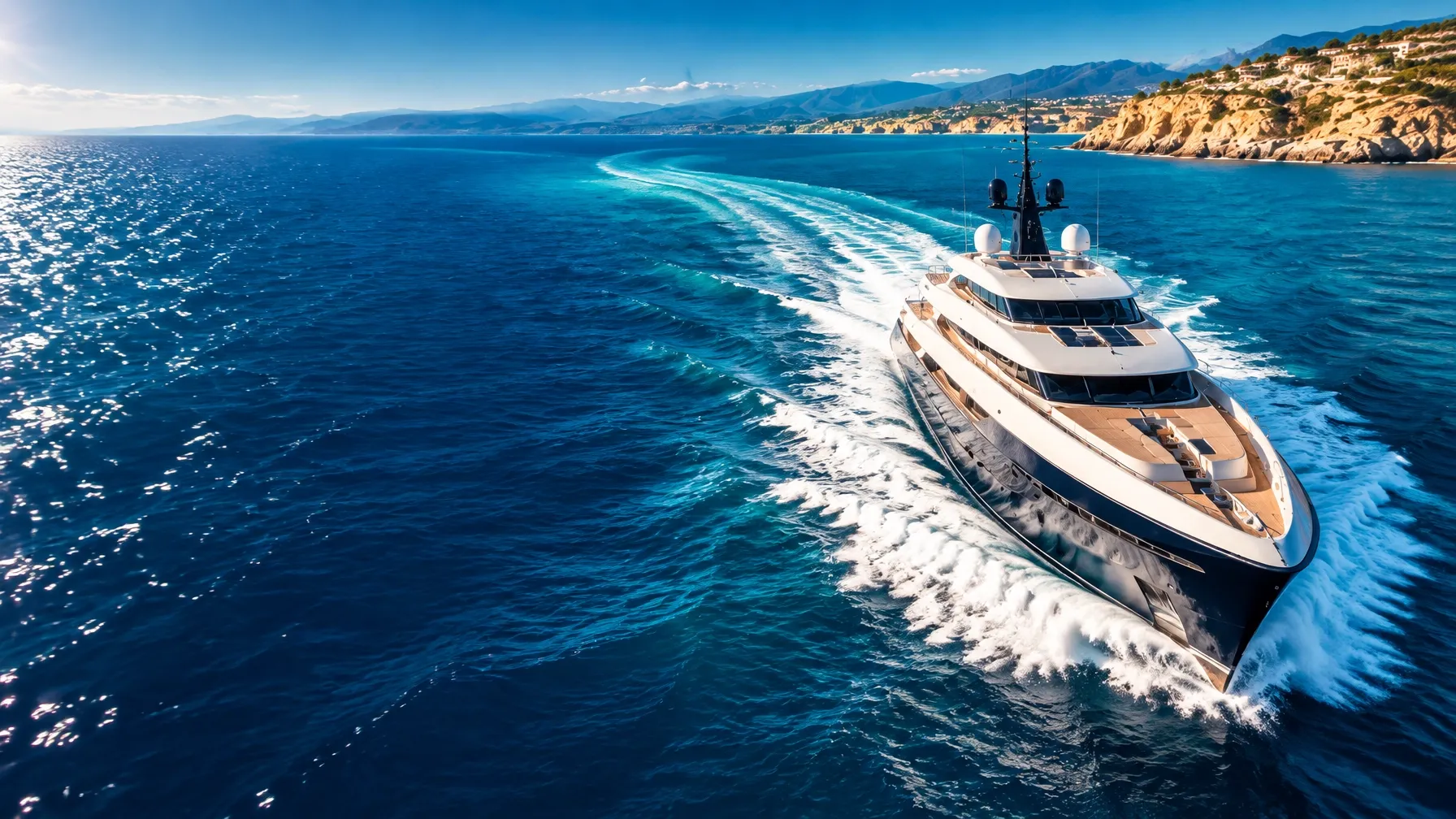A luxury superyacht cuts through brilliant blue water along a sunlit coast